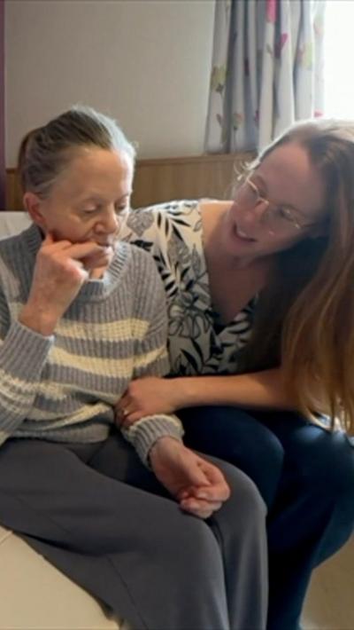 A woman lying in a care home bed with her daughter, a woman with a black and white patterned top and long red hair, by her side