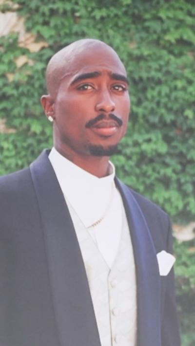 Tupac wears a black suit, white shirt and white vest as he looks towards the camera with greenery behind him.
