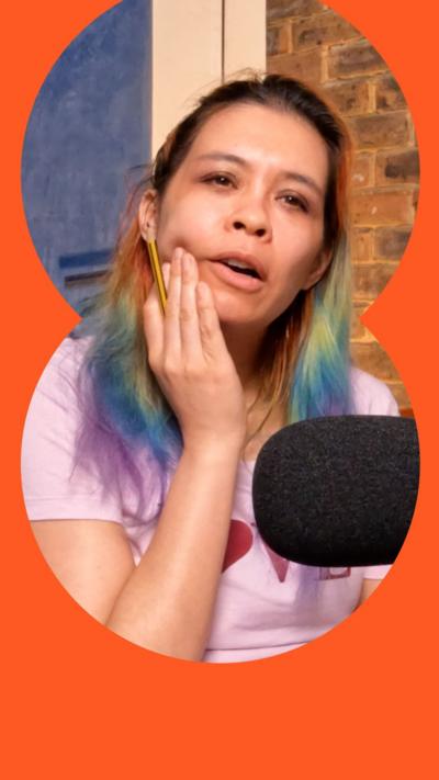 Ria Lina holds her chin while thinking, She has rainbow coloured hair, tied back.