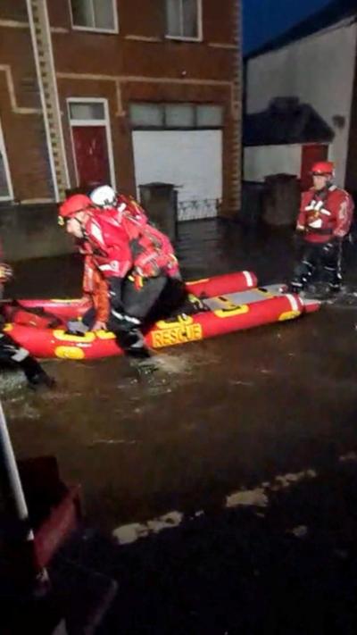 Firefighters dressed in red rescue gear pushing a red dinghy up a flooded street. 