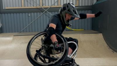A side view of a teenager in a wheelchair, performing a stunt at a skate park, wearing a black helmet.