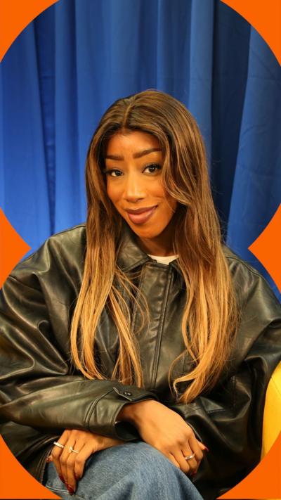 A woman, Kamille, smiles at the camera, She has long brown hair and wears a black leather jacket.