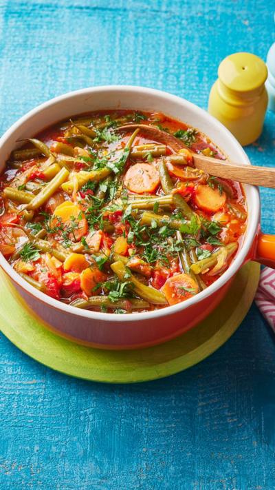 A bowl containing vegetables in a broth with a wooden spoon in, sits on a blue table cloth.