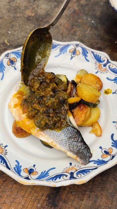 A plate topped with a seabass fillet and roasted potato and courgette, with  thick sauce with capers being dropped on with a spoon.