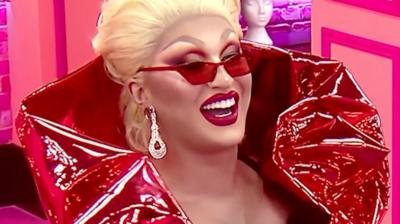 The Vivienne smiles in the Drag Race UK werk room. She wears a red collared outfit, red sunglasses and silver earrings