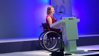 Dr Hannah Barham-Brown presenting on stage in her wheelchair