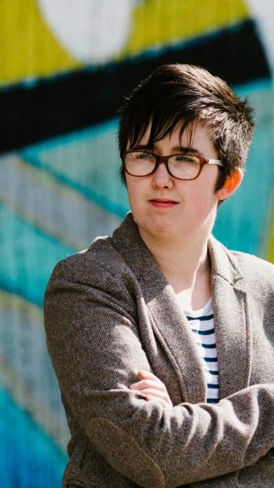 Lyra McKee stands with her arms folded in front of a brightly painted wall. She is a young woman with short black hair, with a fringe over her forehead coming to rest on her glasses. She is smiling She is wearing a brown tweed jacket over a white a navy striped top.