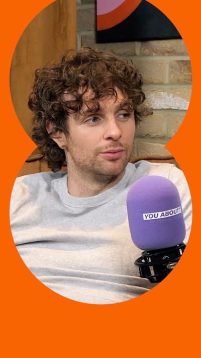 Tom Grennan sits on a couch wearing a grey T-shirt behind a purple microphone