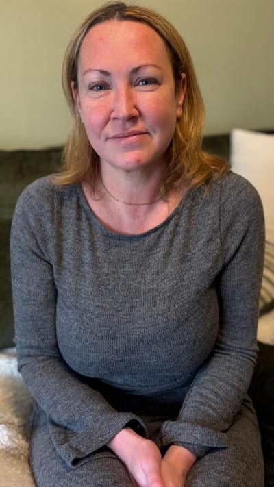 A woman with shoulder-length dark blonde hair. She is staring at the camera and is wearing a grey long-sleeved top. 