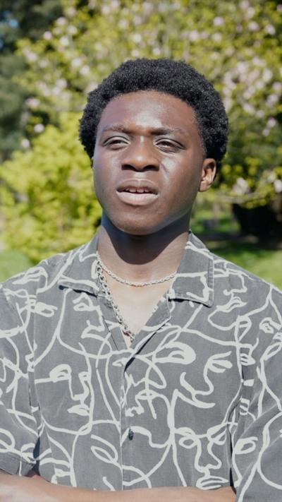 A young black man in a patterned shirt and necklace stood in a park 