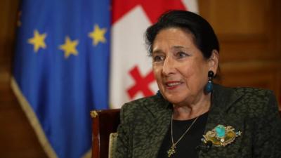 Salome Zourabichvili smiles while sitting in front of Georgia flag and an EU flag