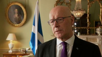 First Minister John Swinney is dressed in a black suit, white shirt and purple tie. He looks slightly to the left of the image. There is a Scottish Saltire in the background along with a gold mirror, gold framed painting and a chandelier reflected in the mirror.