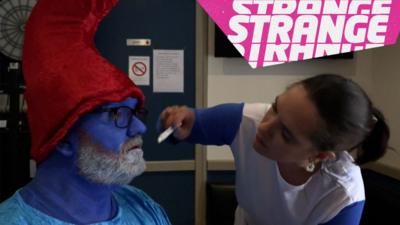 A woman painting the beard of a man dressed as a smurf and the Strange logo