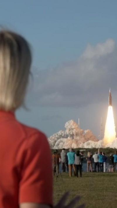 A woman wearing a red top watches from afar as a rocket launches in the distance, with other spectators nearby.