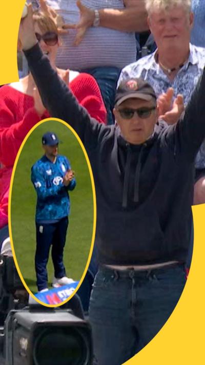 Man holds arms in the air at a cricket stadium with cricket ball in left hand. He is wearing a dark cap and black sunglasses.