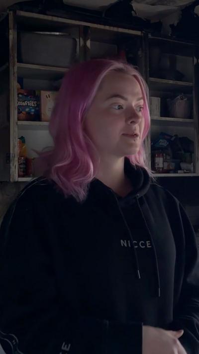 A woman with long pink hair and a black top in a fire damaged kitchen