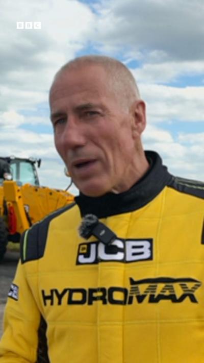 A bald man in a yellow racing suit with the words JCB HYDROMAX on the front