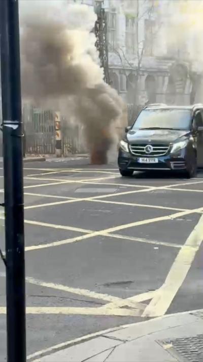 Thick smoke billows from a vent in the middle of a London road.  A London black taxi drives around it.