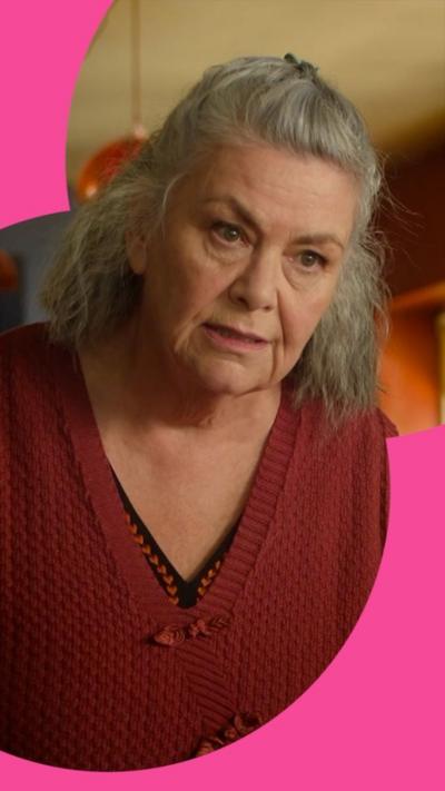 A woman with greay hair cocks her head. She looks serious and wears a red cardigan