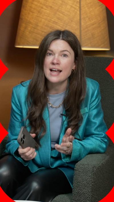 A woman with long brown hair holds a mobile phone. She is wearing a shiny turquoise jacket and a light blue T-shirt.