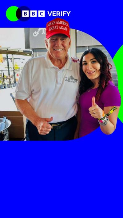 Donald Trump with Laura Loomer. He is wearing a white shirt with a red MAGA hat. She is wearing a t-shirt and giving a thumbs up to the camera.