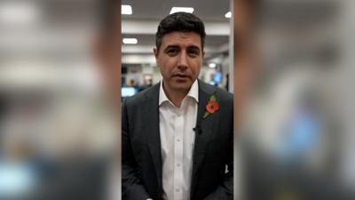 Paris Gourtsoyannis in a white shirt and black blazer. He is wearing a poppy on his left jacket lapel.