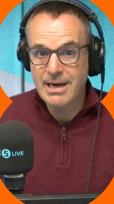 Martin Lewis is wearing a dark red quater-zip jumper. He has radio headphones on and glasses.
