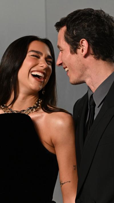 A man and woman laughing together. The woman has long black hair and a black dress. The man has black hair, black suit, black tie and grey shirt