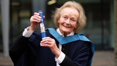 A woman holding a diploma, she is smiling at the camera. She has light hair.