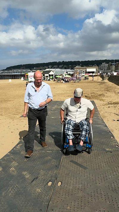 A man walks alongside a man in a wheelchair, both crossing a beach over black rubber mats