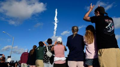Spectators with their back to the camera as the rocket leaves a trail of smoke in the background against a blue sky