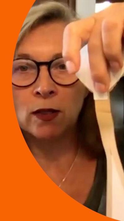A woman with black glasses and grey hair holds up a piece of material to camera