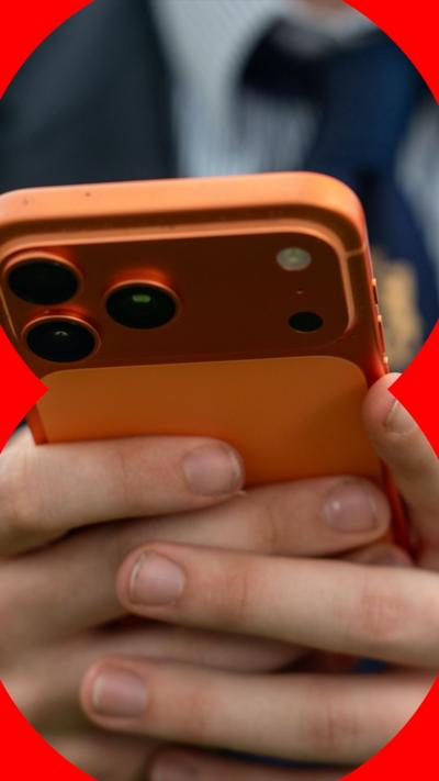 A close-up of a pupil's hands holding a metallic orange mobile phone. They are wearing a school blazer, shirt and tie.