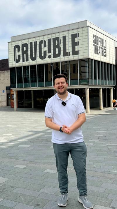 A man wearing a white polo shirt and green trousers stands in front of the Crucible Theatre.
