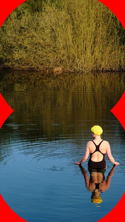 A female wild swimmer stands in a lake by herself wearing a black swimming costume. She wears a yellow swimming cap and has her back to the camera.