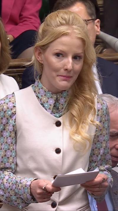 Hannah Spencer speaking in parliament, she is holding a piece of paper in her hands and is wearing a pink waistcoat and patterned shirt