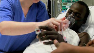 Man lies in hospital bed with drips and oxygen mask