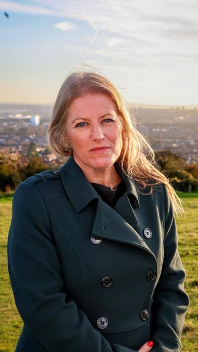 Donna Jones standing on a hill in front of a city. She has long blonde hair and is smiling. She is wearing a green buttoned coat.