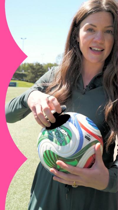 A woman with brown hair lifts a circular cutout from a football to reveal the inside.