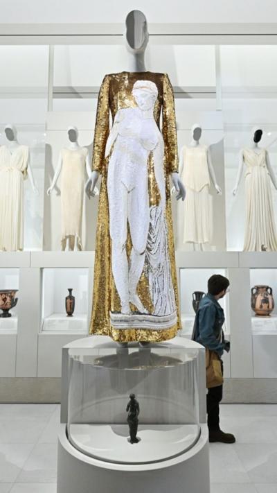 A gold gown stands on a mannequin in front of several white gowns placed on mannequins on shelves.
