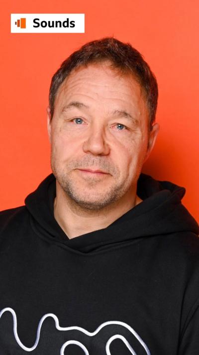 Stephen Graham wearing a black hoodie standing in front of a red background