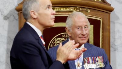 King Charles III sits on a throne while looking at the camera, Mark Carney stands in front of him clapping.
