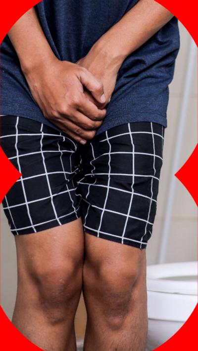 A man holds his hands over his crotch whilst standing next to a toilet. He wears a navy top and checked shorts.