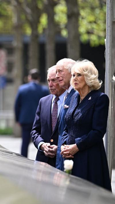 The Queen, King and former New York City mayor Michael Bloomberg stand at the edge of the 9/11 Memorial, looking upwards.
