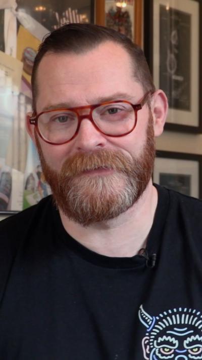 Alex Young looks at the camera. He has short ginger hair with a ginger and white beard. He wears aviator style thick tortoisehell framed glasses and a black t shirt, which has a logo of a viking on it.
