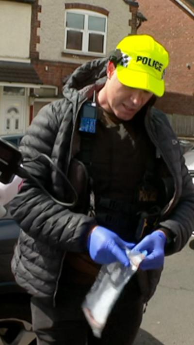 A police officer wearing a black coat and lime green police hat opens a plastic bag wearing blue gloves