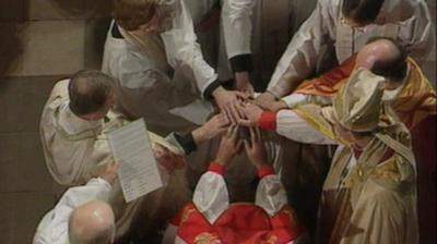 A female priest is ordained in a circle of outreached hands.