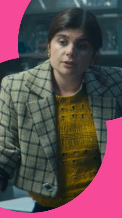 Character Janie stands in an office wearing a grey checked blazer over a yellow top 