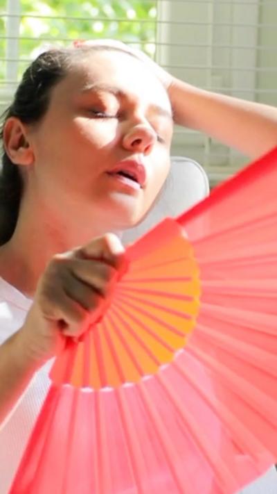 A woman with one hand on her head and her other holding a pink fan. She is sat on white chair in front of a window with white Venetian blinds. 
