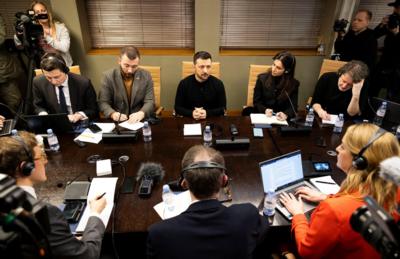 Zelensky sits at a table surrounded by journalists wearing headphones and typing on laptops